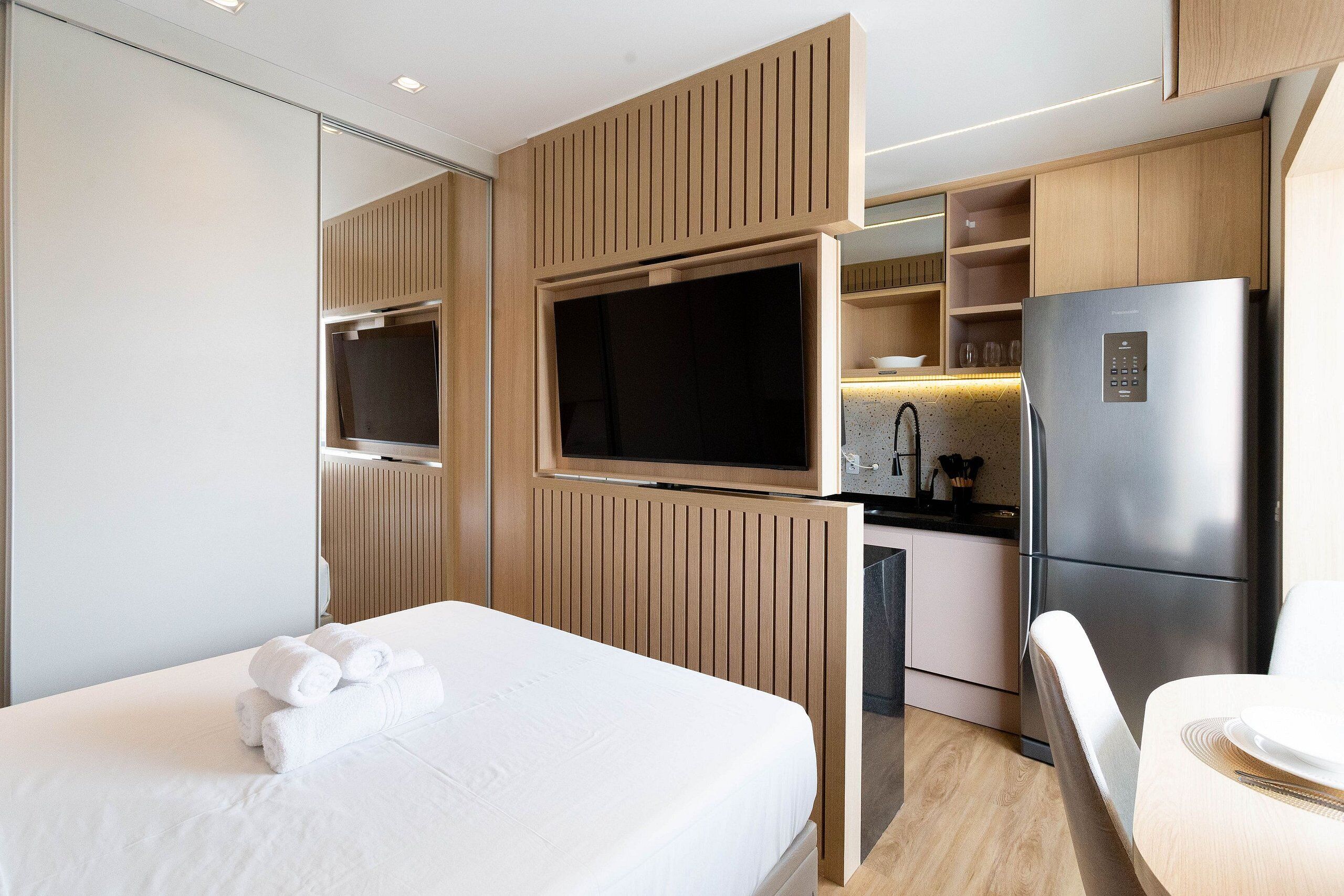 A modern hotel room with a bed, a TV mounted on a wooden panel, a refrigerator, and a small kitchen area.