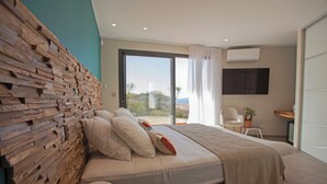 A modern bedroom with a large bed, a wooden headboard, a view of the outdoors, a small desk, and a chair. - Villa Clémentines Porticcio (Grosseto-Prugna)