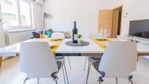 A modern dining area with a white table, four chairs, a bottle of wine, and a TV on the right. - Holiday flat with air conditioning (Pula)