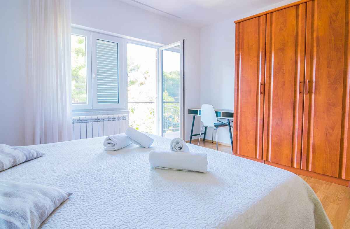 A bedroom with a large bed, a wooden wardrobe, a desk with a chair, and a window with blinds.