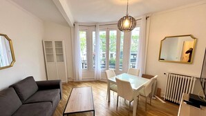 Una sala de estar con sofá, una mesa de centro, una mesa de comedor con sillas, una lámpara colgante, un radiador y una ventana con cortinas.