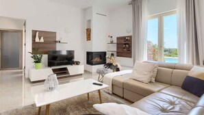 A modern living room with a sofa, TV, fireplace, and large windows. - Villa with pool, air conditioning, gym and parking (Vodnjan)