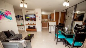 Una sala de estar moderna con un sofá gris, una mesa de comedor de vidrio y sillas color turquesa, y una cocina al fondo.