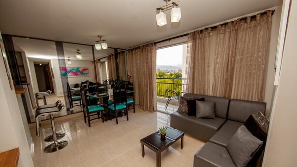Un salón moderno con sofá, mesa de centro y sillas. Cuenta con un balcón con vistas a zonas verdes y edificios.