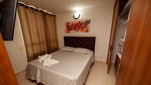 Una cama bien hecha con cubrecama y almohadas blancas, un cabecero con una lámpara circular y un cuadro abstracto y colorido en la pared.