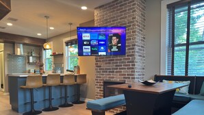 Un área de bar moderna con un televisor mostrando servicios de transmisión, una pared de ladrillo y vistas al exterior.