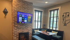 Una habitación acogedora con chimenea de ladrillo, un televisor montado en la pared y una zona de estar con un banco y una mesa.
