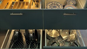 Una cocina con encimera de mármol, sillas de madera y una mesa de comedor. Se observan dos cajones con utensilios y utensilios de cocina.