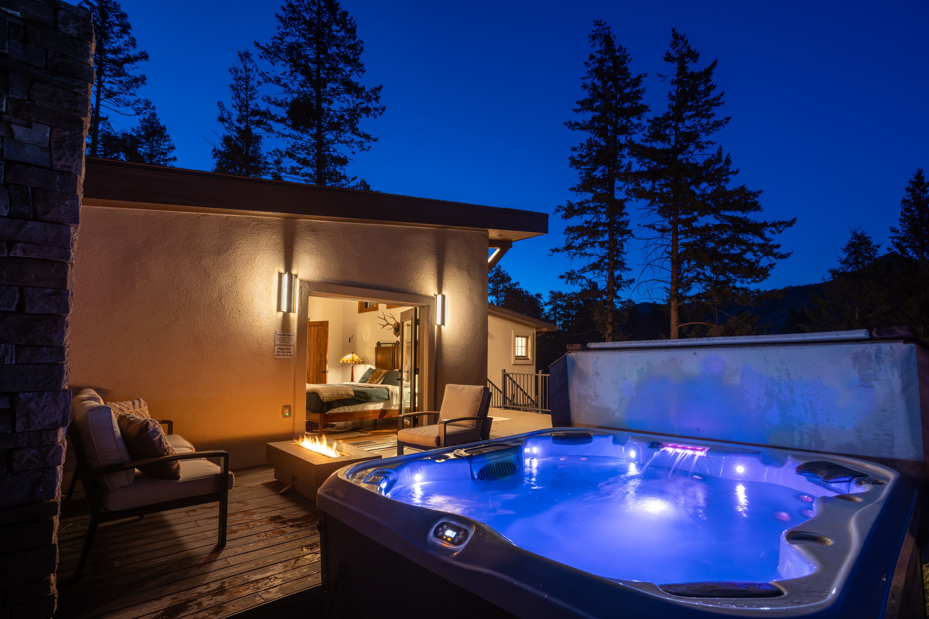 A hot tub on a wooden deck with a stone wall and a bed visible inside a house.