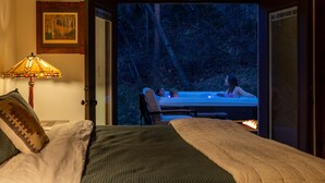 A bedroom with a bed, a nightstand, a lamp, and a view of a hot tub outside. - Serene Mountain View Cabin w/ Deck, Hot Tub, Fire Pit, Forest & Peak Retreats (Cascade-Chipita Park)