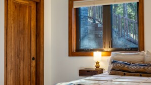 A bedroom with a wooden door, a bed with a patterned bedspread, a nightstand with a lamp, and a window with a view of a wooden deck and trees. - Serene Mountain View Cabin w/ Deck, Hot Tub, Fire Pit, Forest & Peak Retreats (Cascade-Chipita Park)