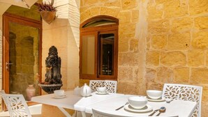 Un espacio de comedor al aire libre con paredes de piedra, mobiliario blanco y una fuente decorativa.