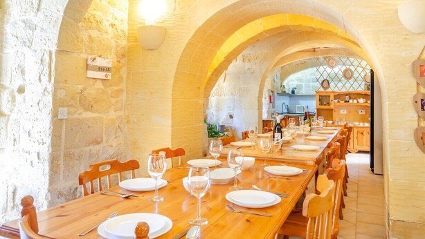 Un comedor con una larga mesa de madera dispuesta para una comida, paredes de piedra y vista hacia una cocina.