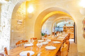 A dining area with a long wooden table set for a meal, stone walls, and a view into a kitchen area. - 300y/o Gozo Country Villa: pool, games, sleeps 14 (Sannat)