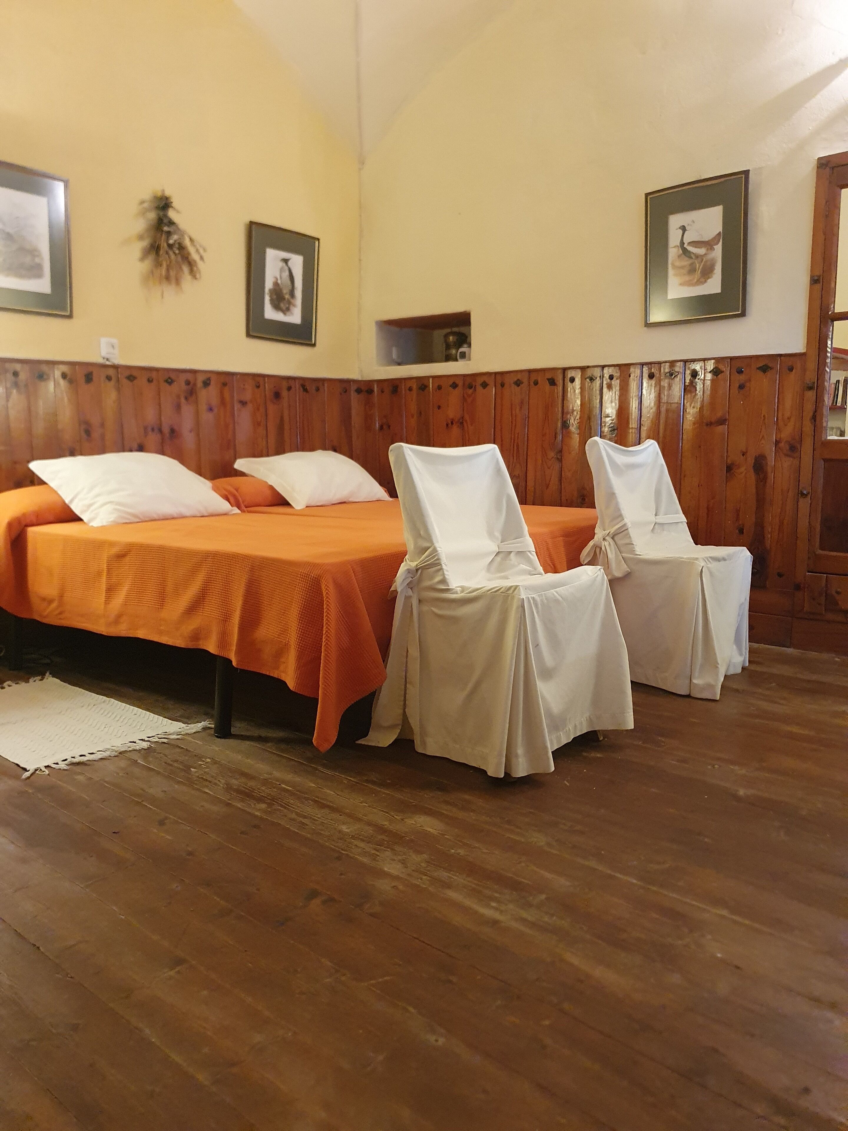 A room with two beds, each with an orange bedspread and white pillows, and two chairs covered with white cloths.