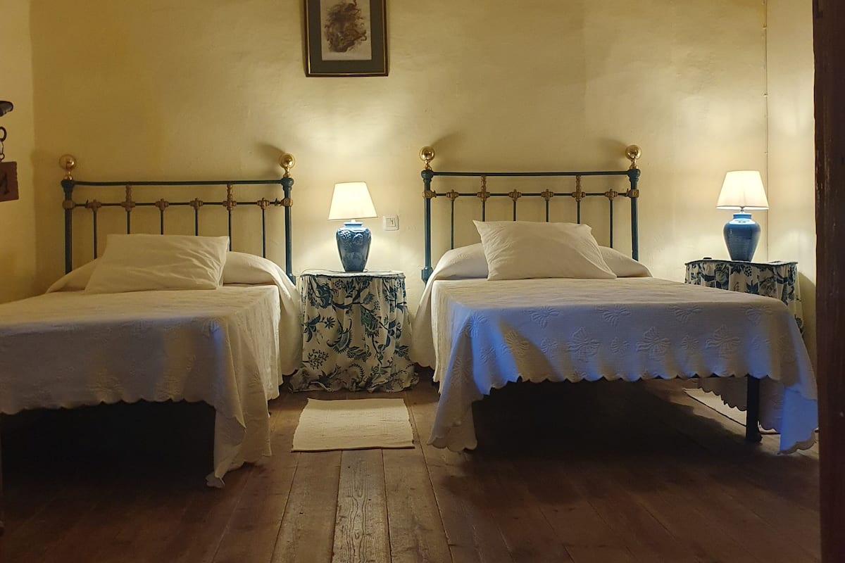 Una habitación con dos camas, cada una con una colcha blanca y una mesita de noche a su lado. Hay un cuadro enmarcado en la pared sobre las camas.