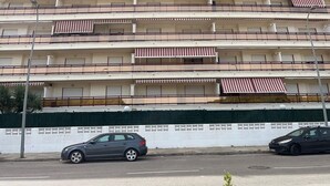 Un edificio de varios pisos con balcones, fachada blanca y una cerca verde al fondo.