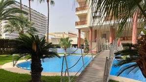 Un área de piscina con muelle de madera, palmeras y un edificio con balcones al fondo.