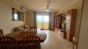 Un área de comedor con una mesa y sillas de madera, un televisor sobre un mueble de madera y un balcón con vistas.