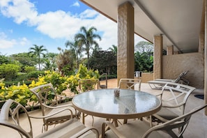Une terrasse extérieure avec une table ronde en verre, entourée de chaises blanches, le tout se fondant dans un décor luxuriant de verdure et de palmiers.