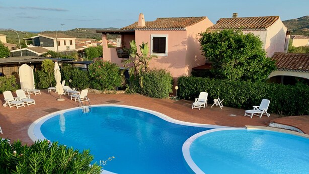 Una piscina con sillas reclinables blancas y una pérgola, rodeada de vegetación y arquitectura tradicional.