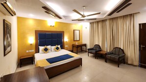 A hotel room with a large bed, a desk, two chairs, a ceiling fan, and a large window with curtains. - Design galleries complex (Faridabad)