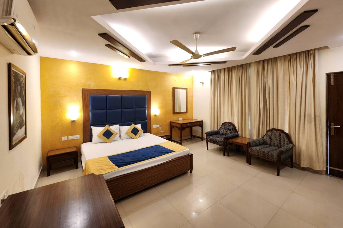 A hotel room with a large bed, a desk, two chairs, a ceiling fan, and a large window with curtains.