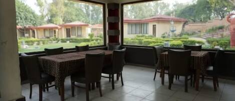 Un espace repas avec des tables et des chaises, de grandes fenêtres donnant sur un jardin et des bâtiments.