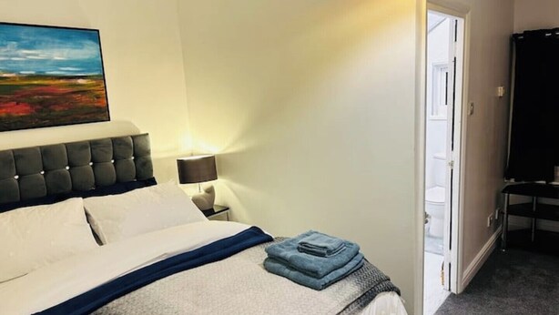 A neatly made bed with a blue blanket and pillows, a painting on the wall, and a door leading to a balcony. - 1 Luxe Exec Bedroom Apt Derby (Derby)