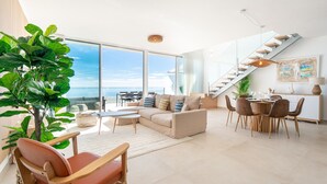 Amplia sala de estar con una planta grande, un sofá, una mesa de comedor con sillas y una escalera que conduce a un nivel superior.