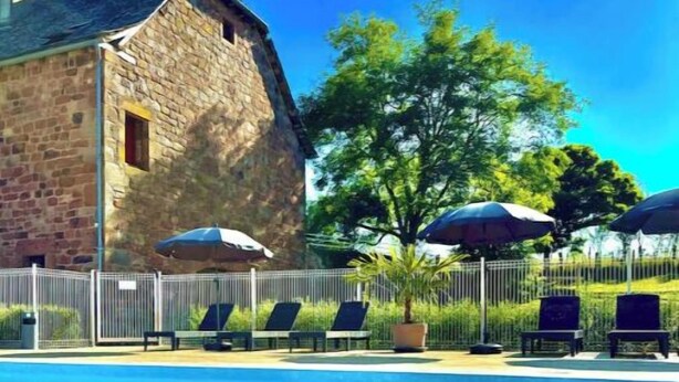 Une piscine avec des chaises longues et des parasols, un bâtiment en pierre et un espace clos avec des arbres.