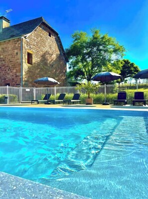 A swimming pool with lounge chairs and umbrellas, a stone building, and a fenced area with trees.