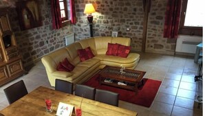 Un salon chaleureux avec un mur en pierre, un canapé beige agrémenté de coussins rouges, une table basse en bois et une table à manger avec des chaises.