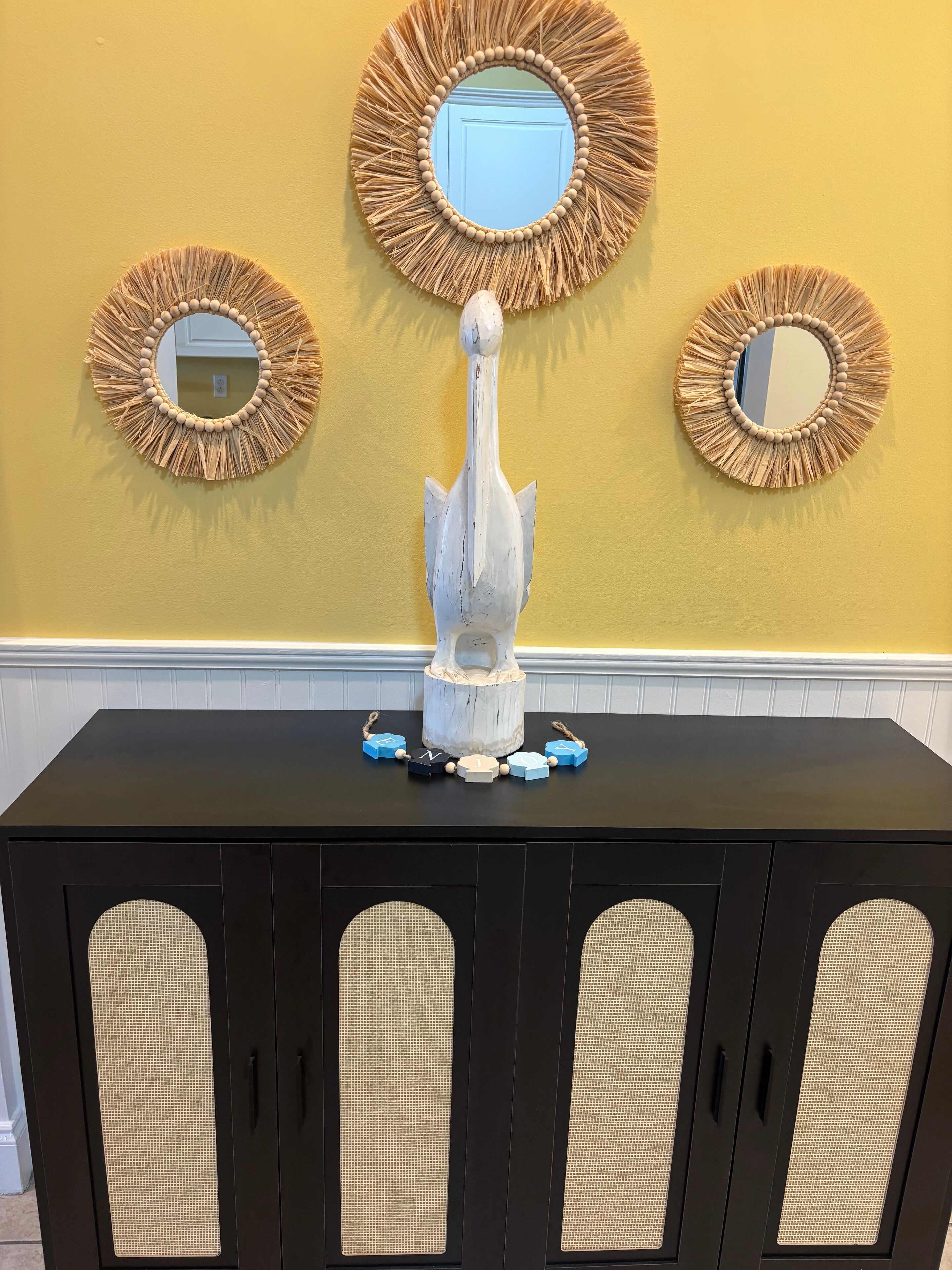 A black console table with a decorative sculpture and a beaded necklace centerpiece. Above the table, there are three wall-mounted mirrors with a woven frame design.
