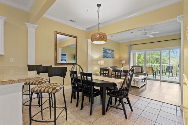 A dining area with a large table, black chairs, and a chandelier. There is a balcony with outdoor furniture. - Family Tides at Litchfield (Pawleys Island)