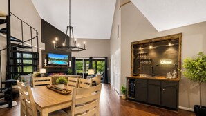 Une cuisine moderne dotée d’un meuble à vin intégré, un coin repas avec des meubles en bois et un grand écran de télévision diffusant un paysage pittoresque.