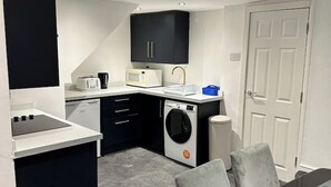 Een compacte keuken met een magnetron, broodrooster en een wasmachine. Er is een eettafel met stoelen en een witte deur op de achtergrond.