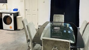 Een moderne eethoek met een glazen tafel, grijs beklede stoelen, een ingebouwde wasmachine en een donker gordijn.