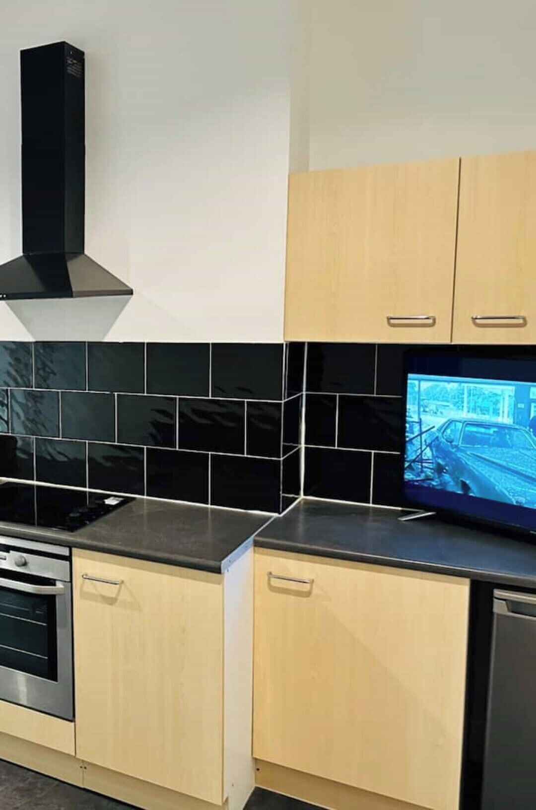 A kitchen with light wood cabinets, black tiles, a black range hood, and a television displaying a car accident.