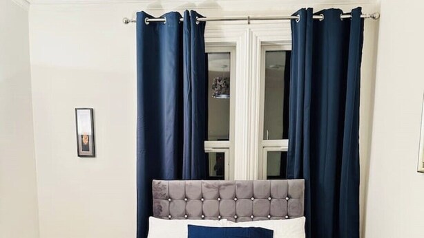 Una cama bien hecha con un cabecero tapizado en gris, un edredón azul y blanco, y una toalla doblada al pie.
