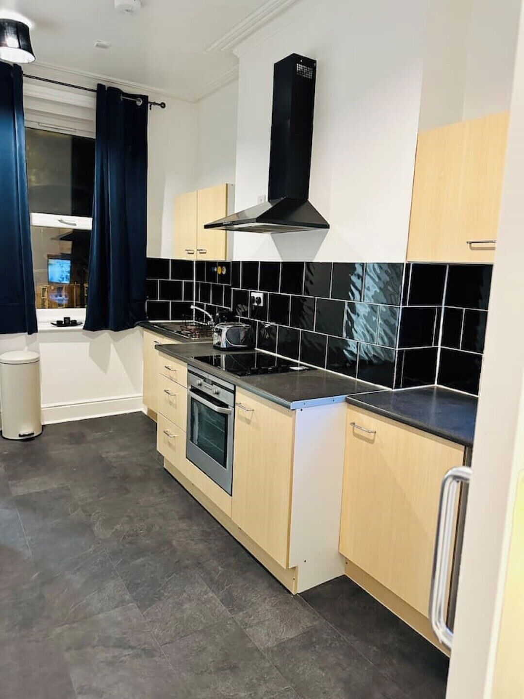 A modern kitchen with a stove, oven, and dishwasher, light wood cabinets, black and white tiles, and a black range hood.