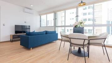 A modern living room with a blue sofa, a TV, a dining table with chairs, and large windows offering a city view.