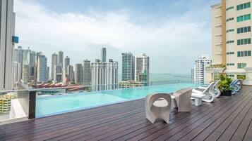 A rooftop pool area with a city view, lounge chairs, and a wooden deck.