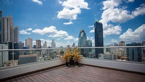 Eine Dachterrasse mit Holzboden und Blumentöpfen, mit Blick auf eine Stadtlandschaft mit modernen Gebäuden und blauem Himmel.