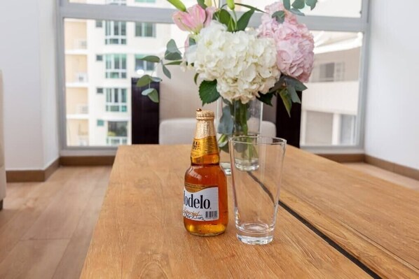 A bottle of Modelo beer and a glass on a wooden table with a vase of flowers in the background.