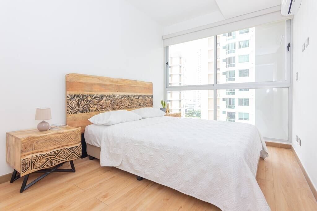 A bedroom with a wooden headboard, a white bedspread, a bedside table with a lamp, and a large window with a city view.