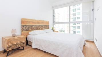 A bedroom with a wooden headboard, a white bedspread, a bedside table with a lamp, and a large window with a city view.