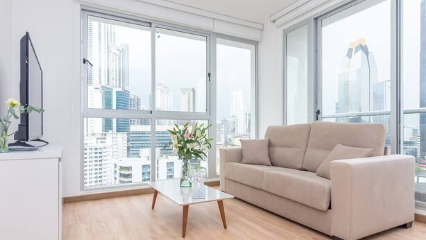 A modern living room with a sofa, a coffee table, and a TV. Large windows offer a city view.