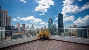 A rooftop terrace with a wooden floor and potted plants, overlooking a city skyline with modern buildings and a clear sky.
