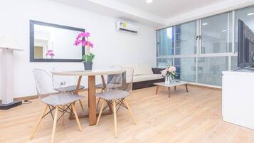 A modern hotel room with a dining area, a sofa, a small table with a vase of flowers, and a large window with a view of the city.